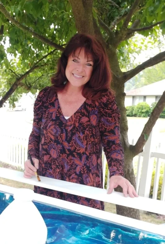 Kristi Moomey smiling under a tree, paintbrush in hand, beside one of her blue paintings